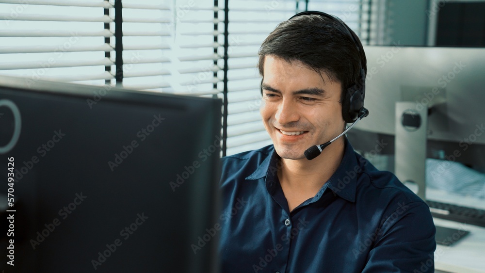 Young competent asian male call center agent working at his computer while simultaneously speaking w