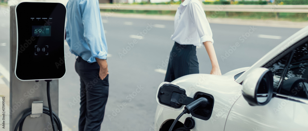 Progressive businessman and businesswoman with electric car connected to charging station before dri