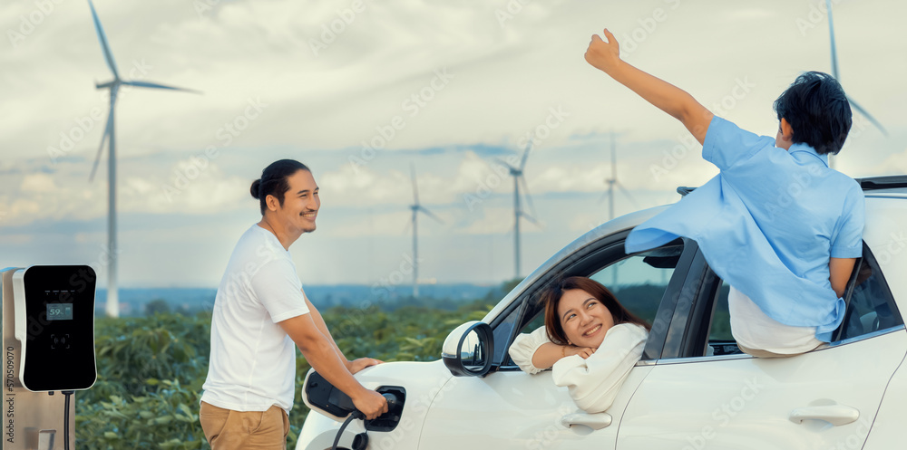 Concept of progressive happy family enjoying their time at wind farm with electric vehicle. Electric