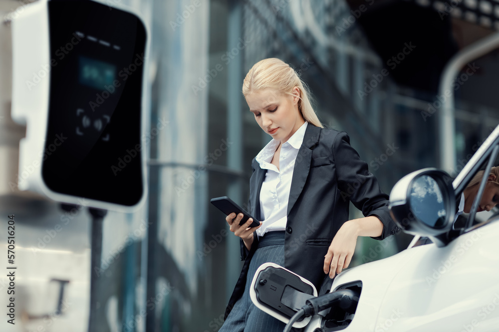 Businesswoman wearing black suit using smartphone, leaning on electric car recharge battery at charg