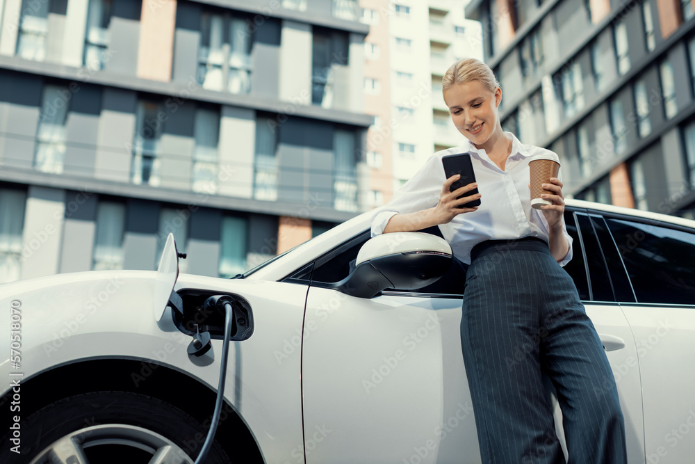 Focus businessman using phone, leaning on electric vehicle, holding coffee with blurred city residen