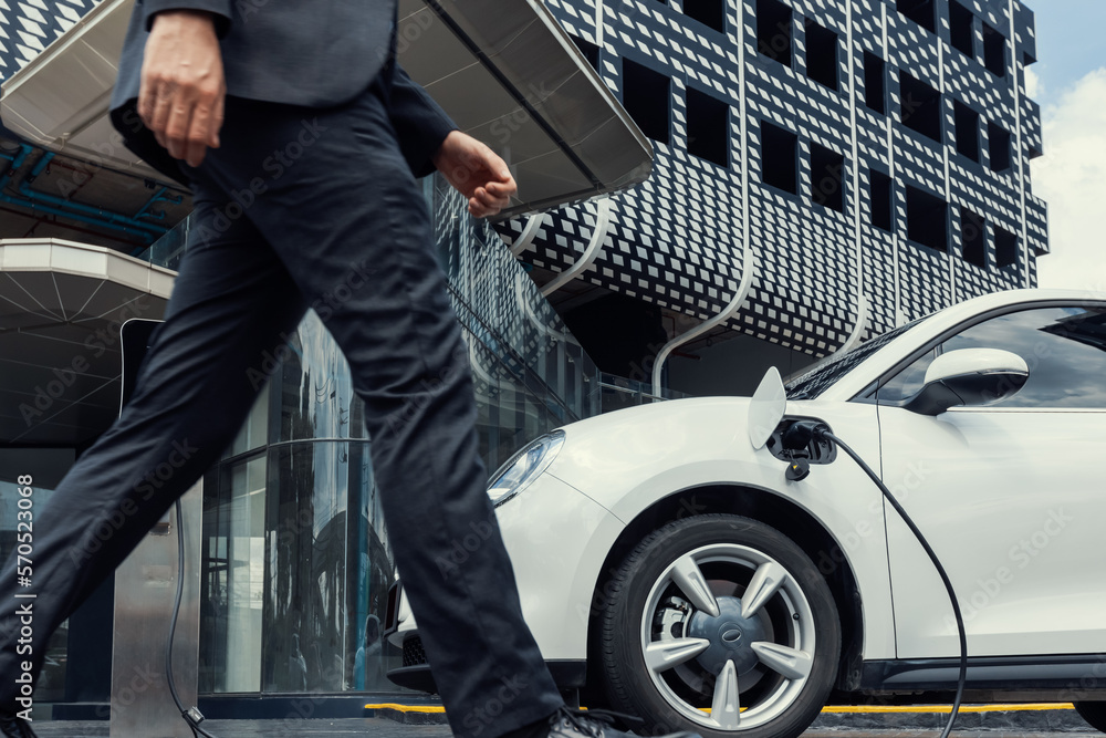 Closeup progressive businessman with electric car recharging at public charging station with backgro