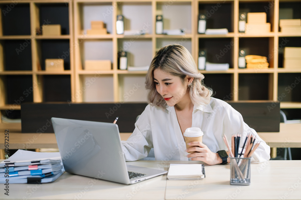 Confident beautiful Asian businesswoman typing laptop computer and digital tablet while holding coff