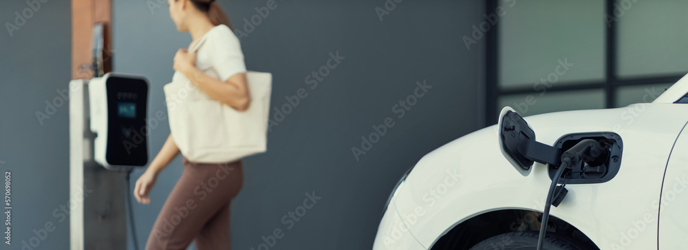 Focus image of electric vehicle recharging battery at home charging station with blurred woman walki