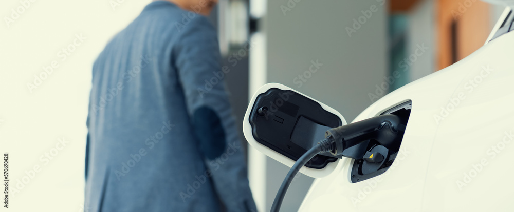 Focus electric car charging at home charging station with blurred progressive man walking in the bac