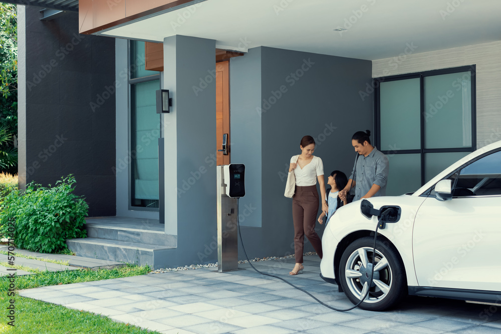 Progressive young parents and daughter with electric vehicle and home charging station. Green and cl