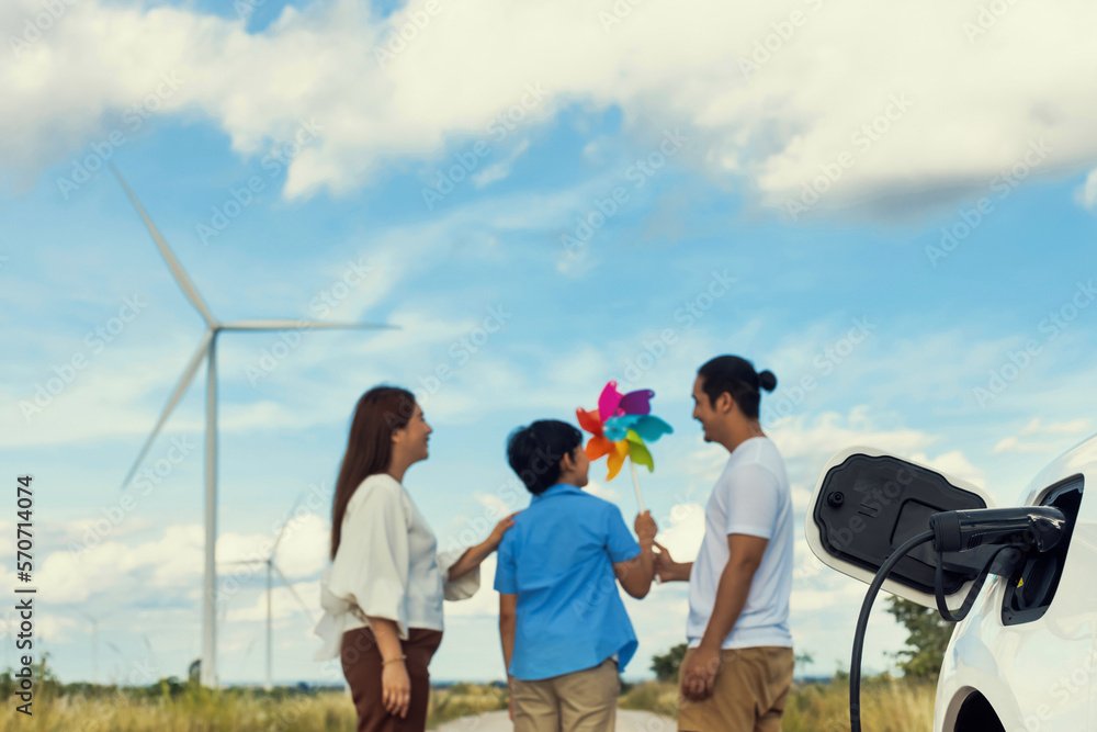 Concept of progressive happy family holding windmill toy and relax at wind farm with electric vehicl