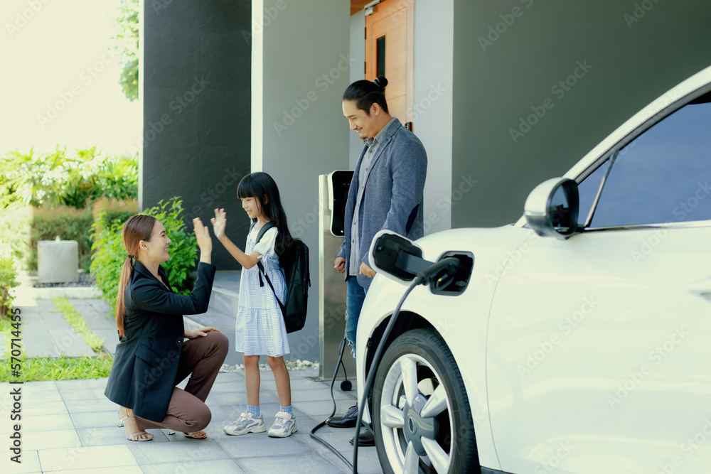 Progressive young parents and daughter with electric vehicle and home charging station. Green and cl