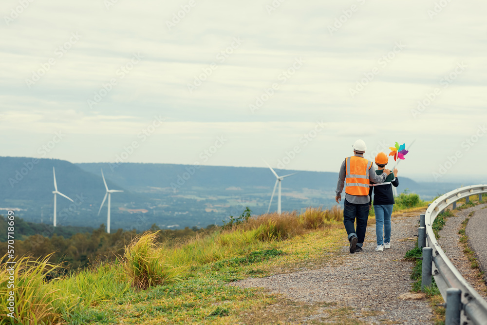 Engineer with his son holding windmill toy on a wind farm atop a hill or mountain. Progressive ideal