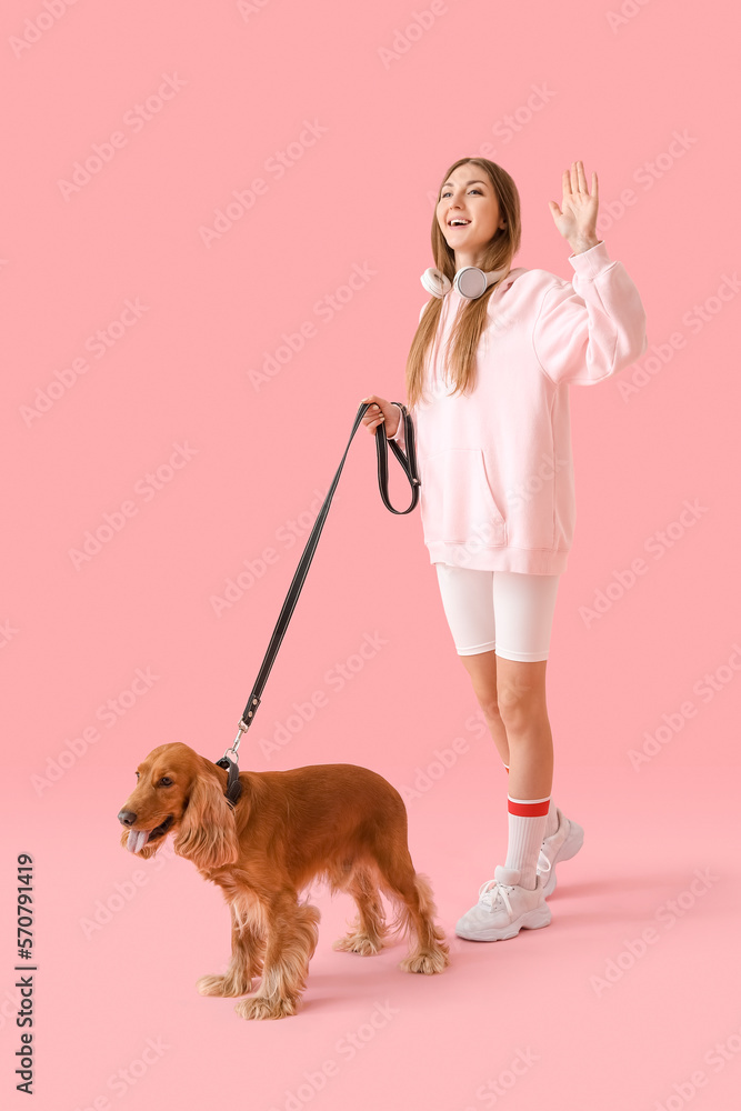 Young woman in headphones with cute cocker spaniel walking on pink background