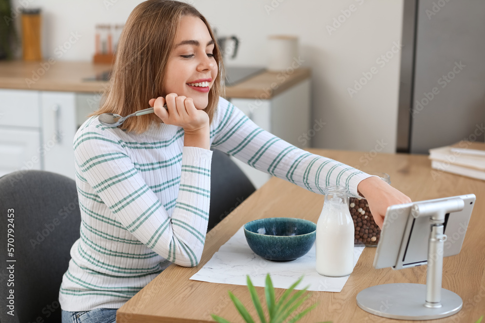 Young woman with spoon using tablet computer at table in kitchen