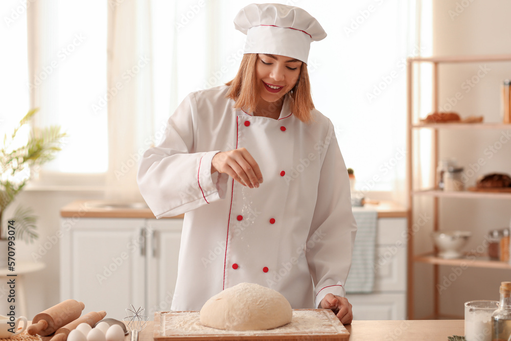 Female baker sprinkling dough with flour at table in kitchen