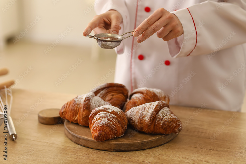 Female baker sprinkling croissants with sugar powder at table in kitchen, closeup