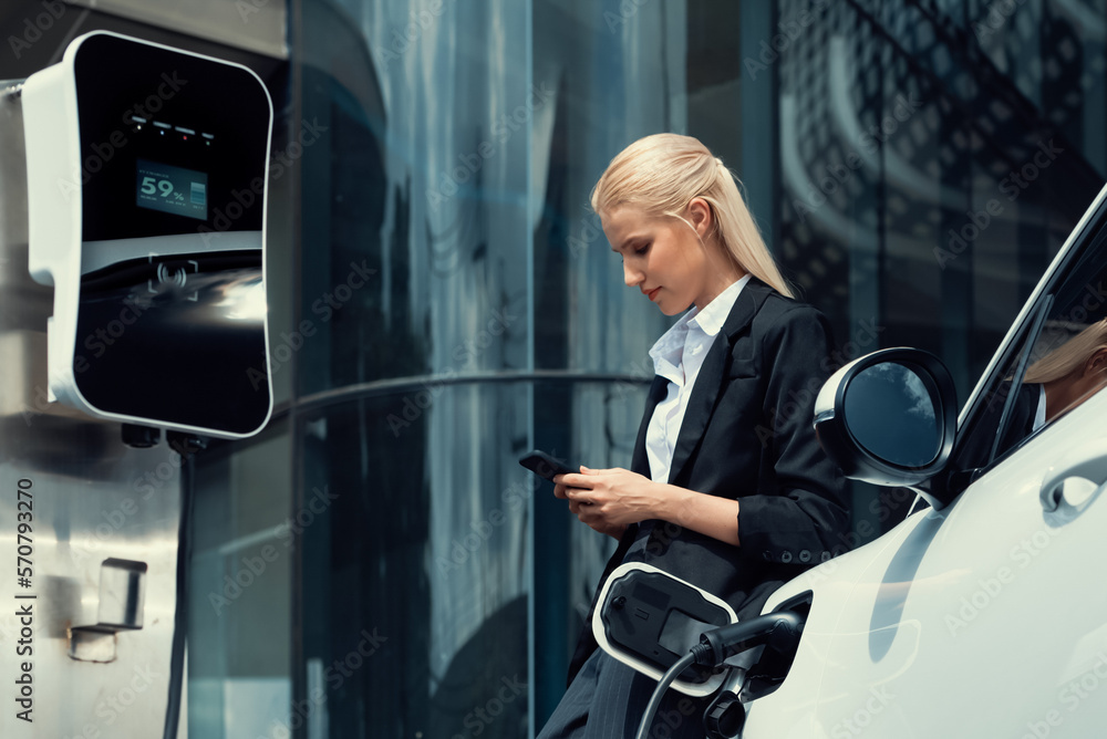 Businesswoman wearing black suit using smartphone, leaning on electric car recharge battery at charg