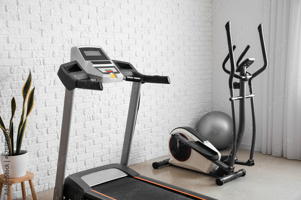 Interior of gym with modern treadmill and elliptical trainer near white brick wall