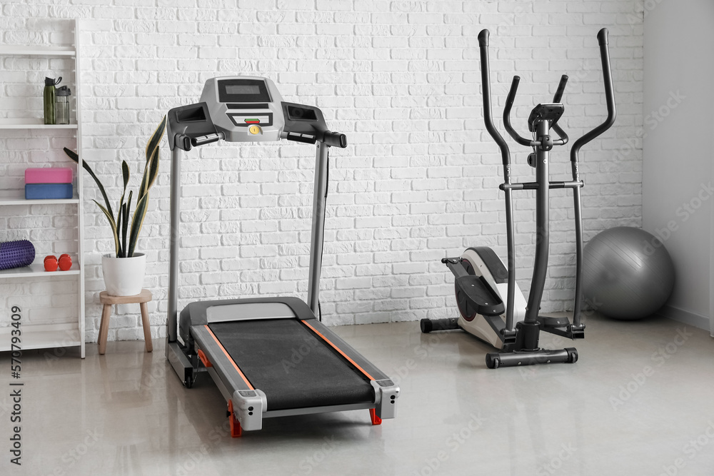 Interior of gym with modern treadmill and elliptical trainer near white brick wall