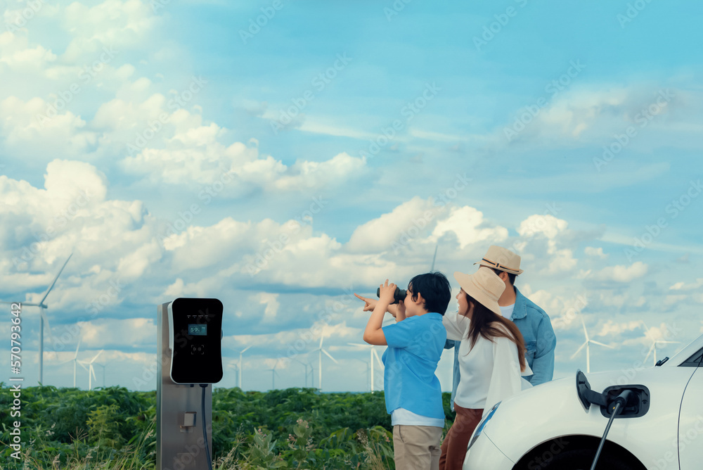 Concept of progressive happy family enjoying their time at wind farm with electric vehicle. Electric