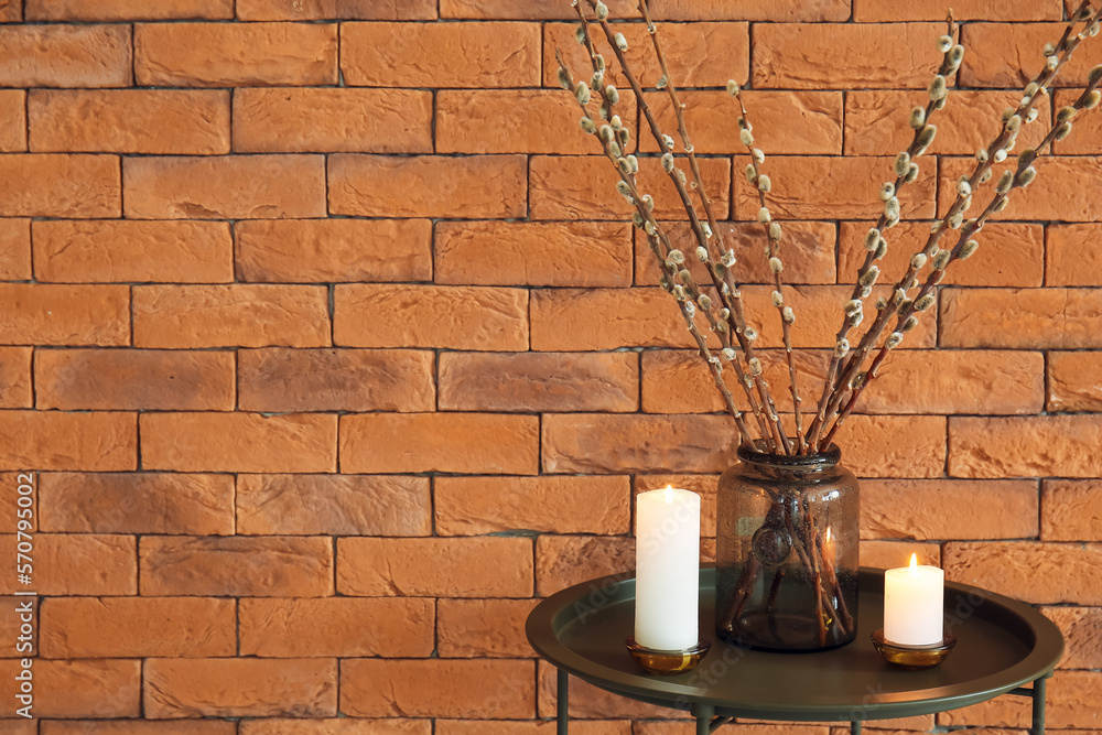 Vase with willow branches and burning candles on table near brick wall