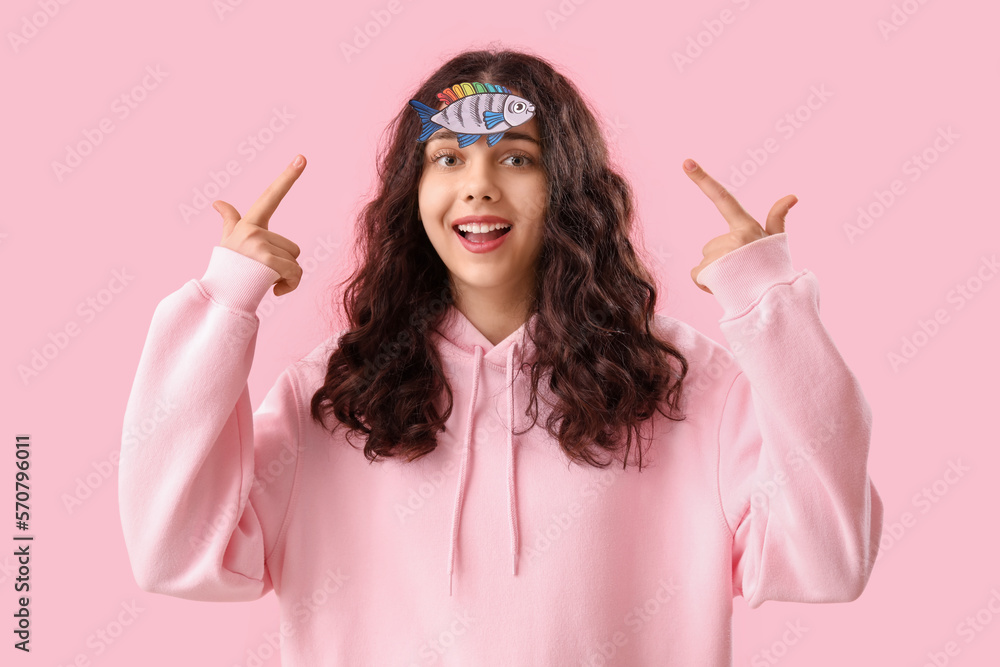 Teenage girl pointing at paper fish on pink background. April Fools Day celebration