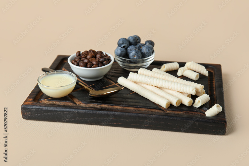 Board with delicious wafer rolls, blueberries, coffee beans and condensed milk on beige background