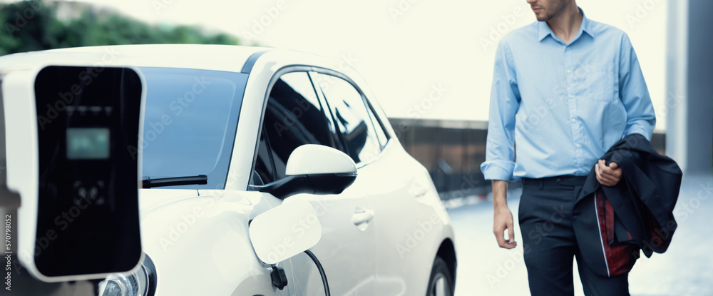 Progressive businessman with electric car recharging at public parking car charging station at moder