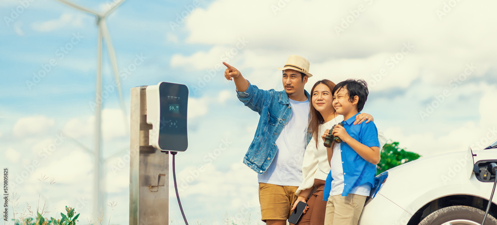 Concept of progressive happy family enjoying their time at wind farm with electric vehicle. Electric