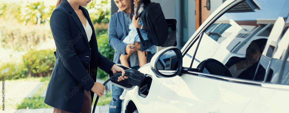 Progressive young parents and daughter with electric vehicle and home charging station. Green and cl