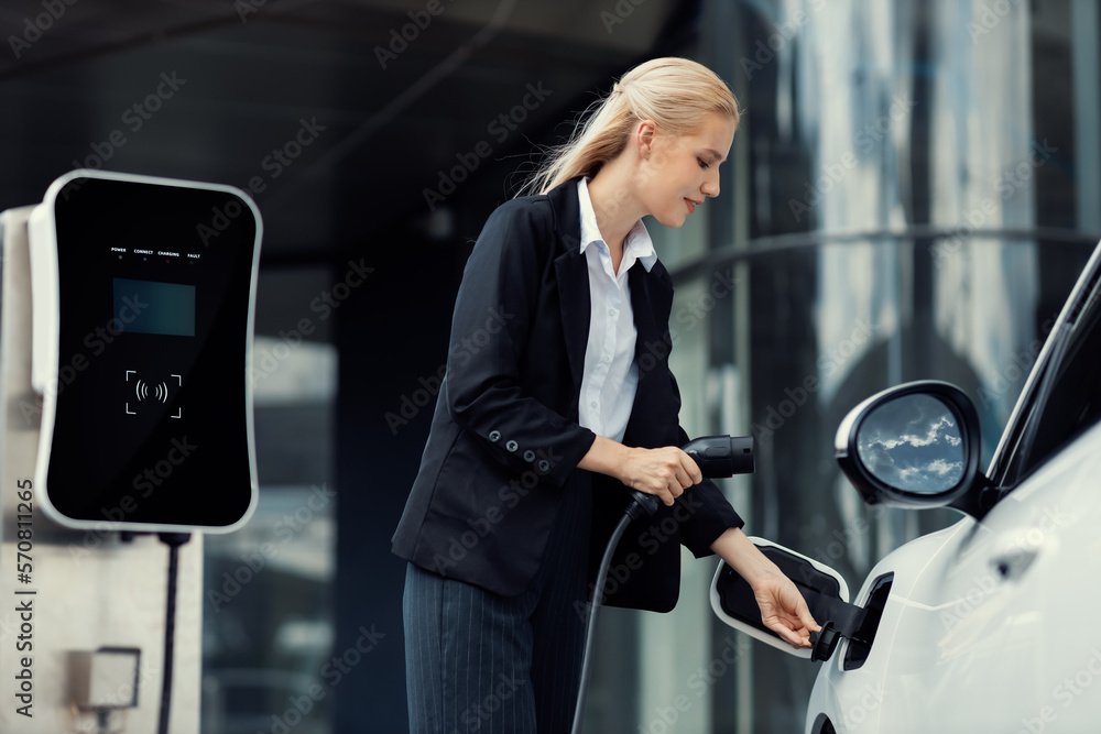 Progressive businesswoman insert charger plug from charging station to her electric vehicle with apa