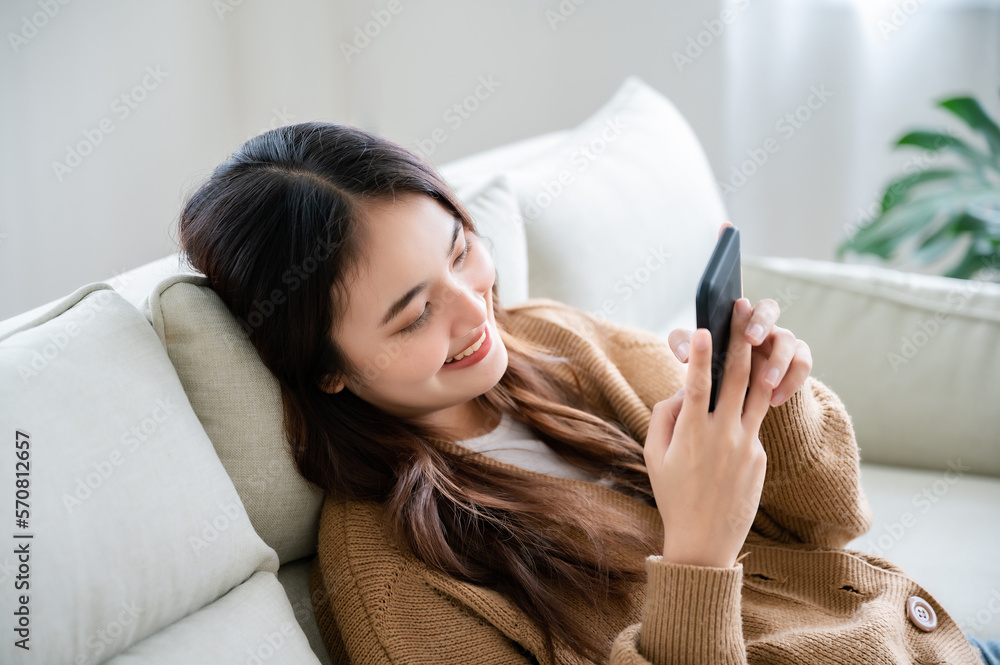 Happy young asian woman relaxing at home. Female is lying down on sofa and using mobile smartphone