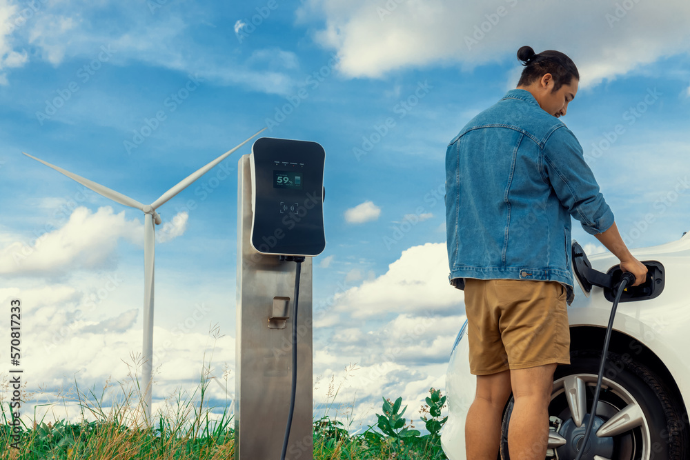 Progressive man with his electric car, EV car recharging energy from charging station on green field