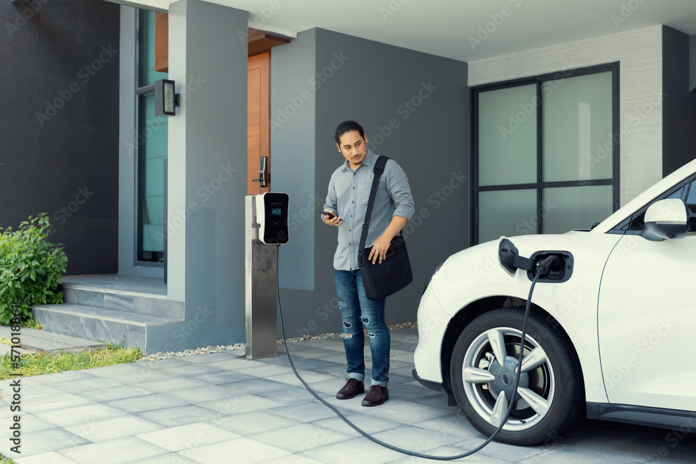 Progressive asian man and electric car with home charging station. Concept of the use of electric ve