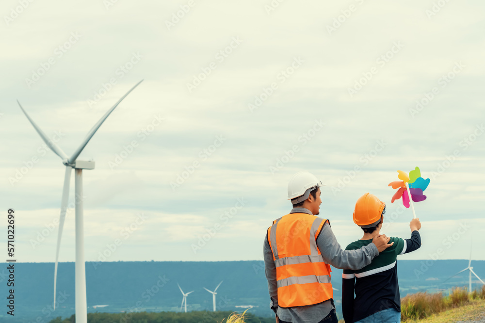 Engineer with his son holding windmill toy on a wind farm atop a hill or mountain. Progressive ideal