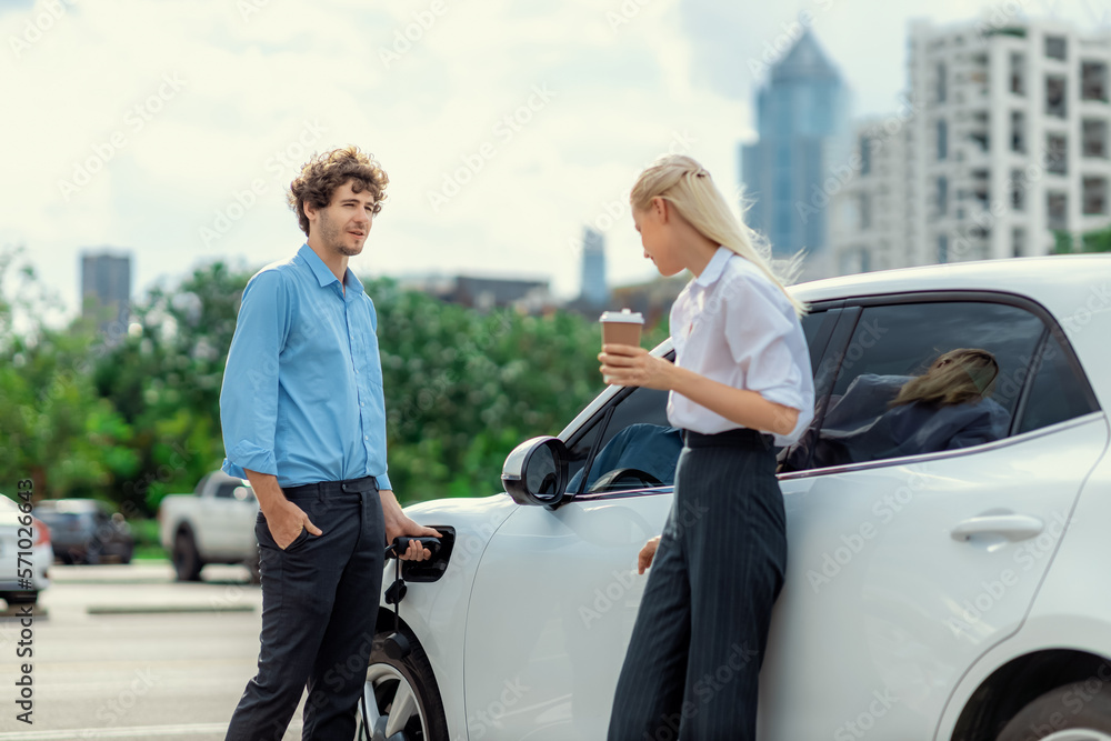 Progressive businessman and businesswoman with coffee, standing at electric car connected to chargin