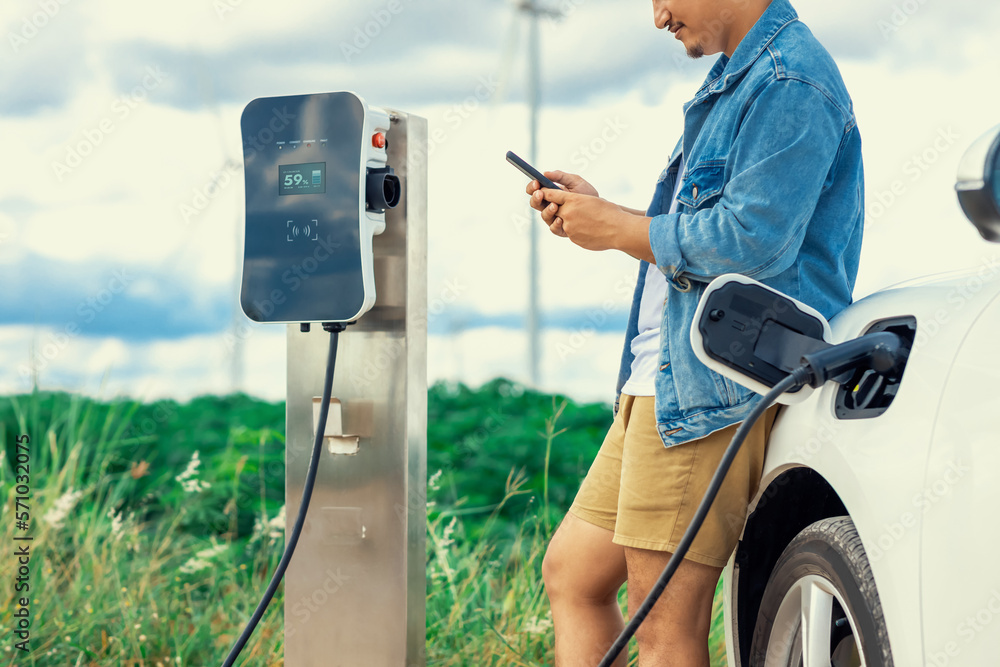 Progressive man with his electric car, EV car recharging energy from charging station on green field