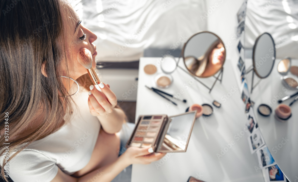 A pregnant woman applies makeup at home in front of a mirror.