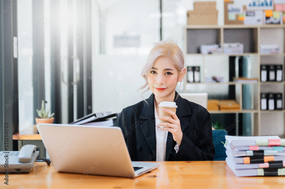 Confident beautiful Asian businesswoman typing laptop computer and digital tablet while holding coff