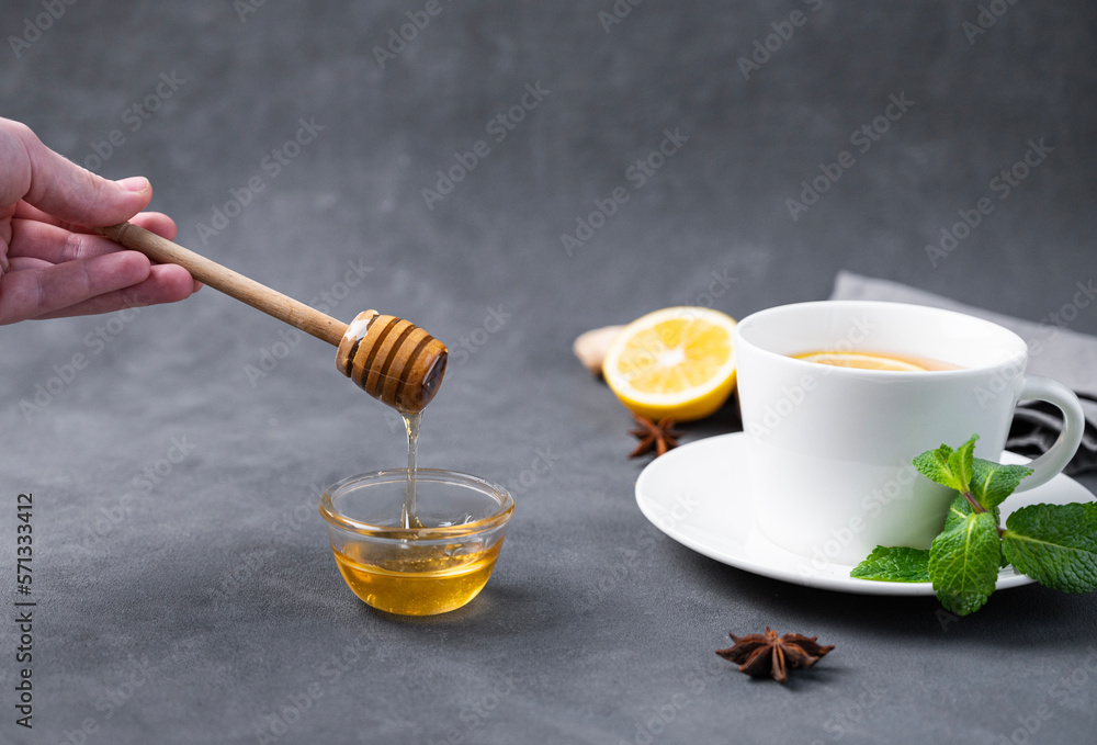 Honey flows from a wooden stick into a glass bowl close-up on a dark background with a cup of tea wi