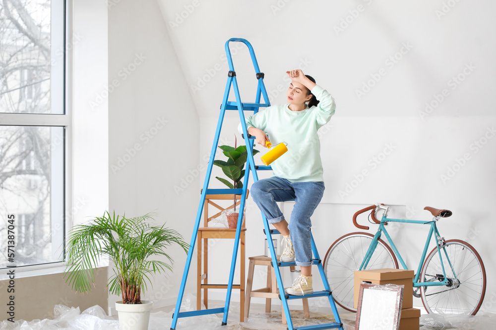 Tired young woman with paint roller and ladder at home