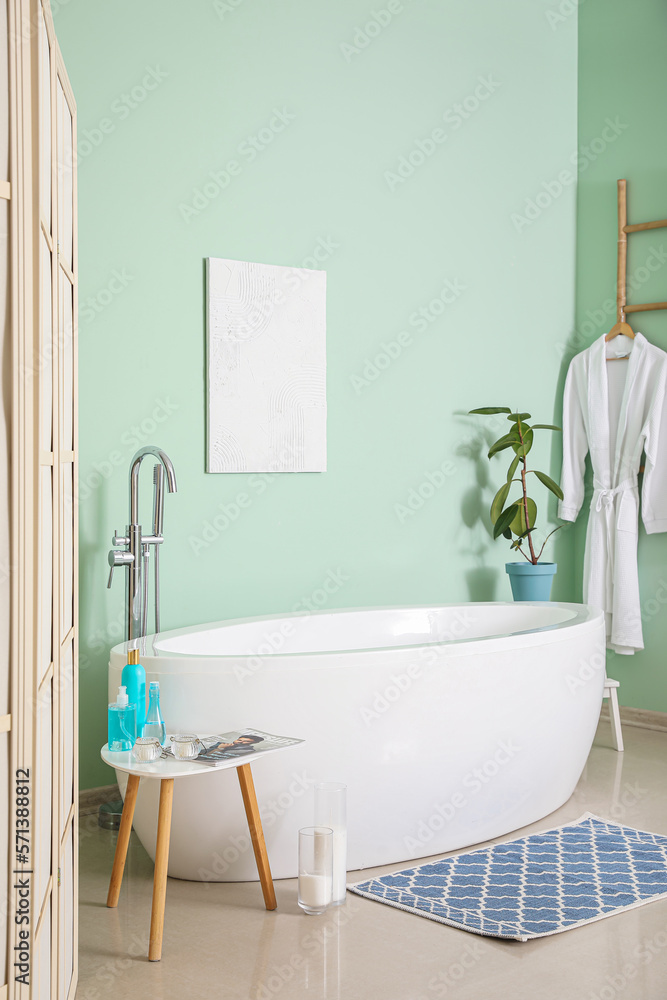 Interior of bathroom with modern bathtub and bath accessories near green wall