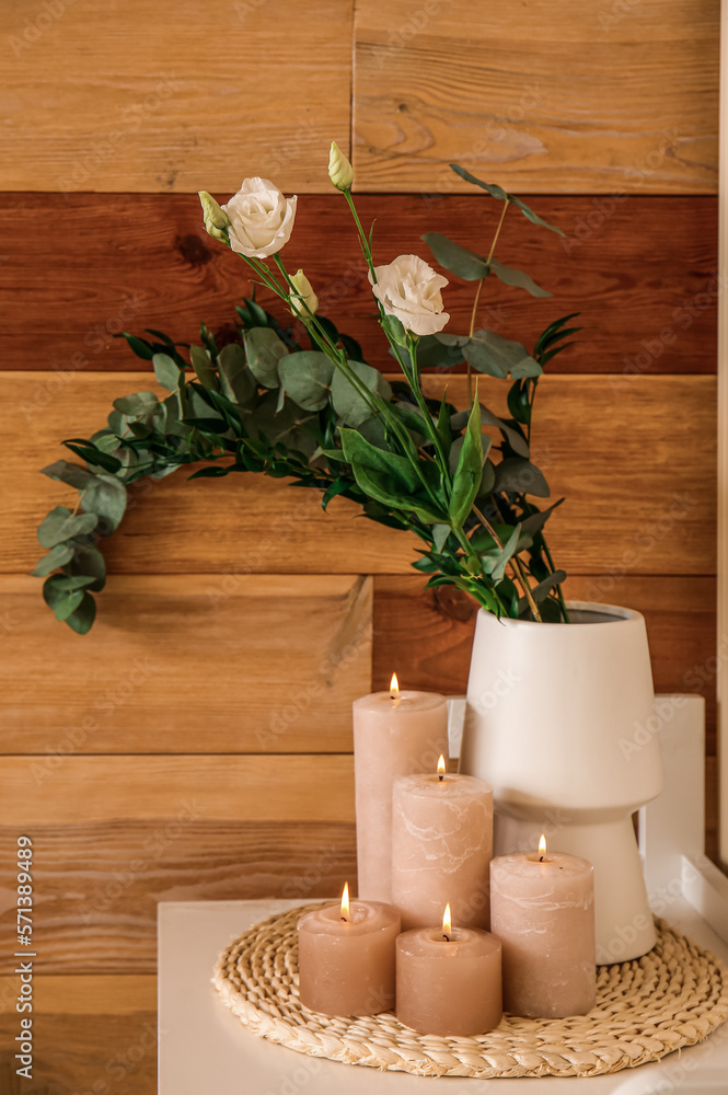 Burning candles, vase with flowers on table near wooden wall