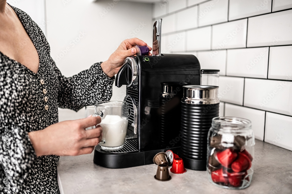 Girl with capsule coffee machine and milk in transparent cup preparing cappuccino at kitchen. Woman 