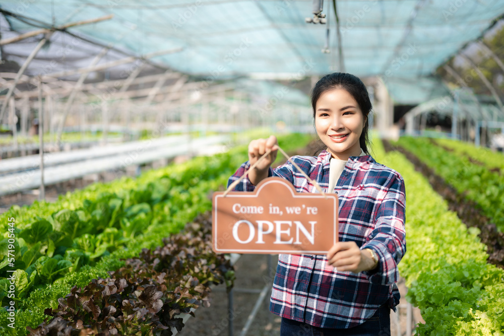  Woman is standing in greenhouse with we are open sign smiling looking at camera welcoming buyers fo