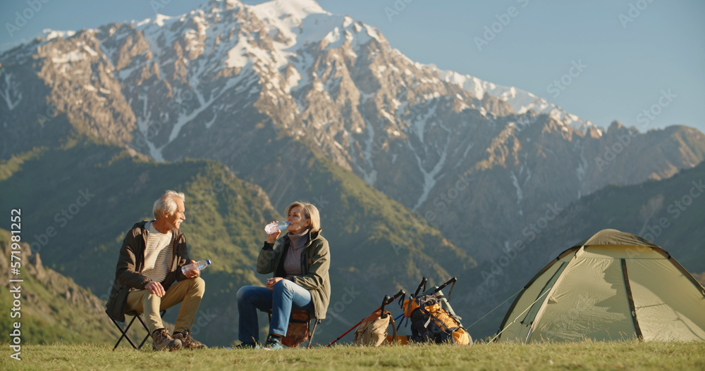 Old caucasian couple taking a rest on top of mountain, sitting and rehydrating while enjoying the vi