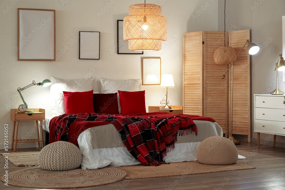 Interior of bedroom with red checkered blankets on bed, blank frames and glowing lamps late in eveni