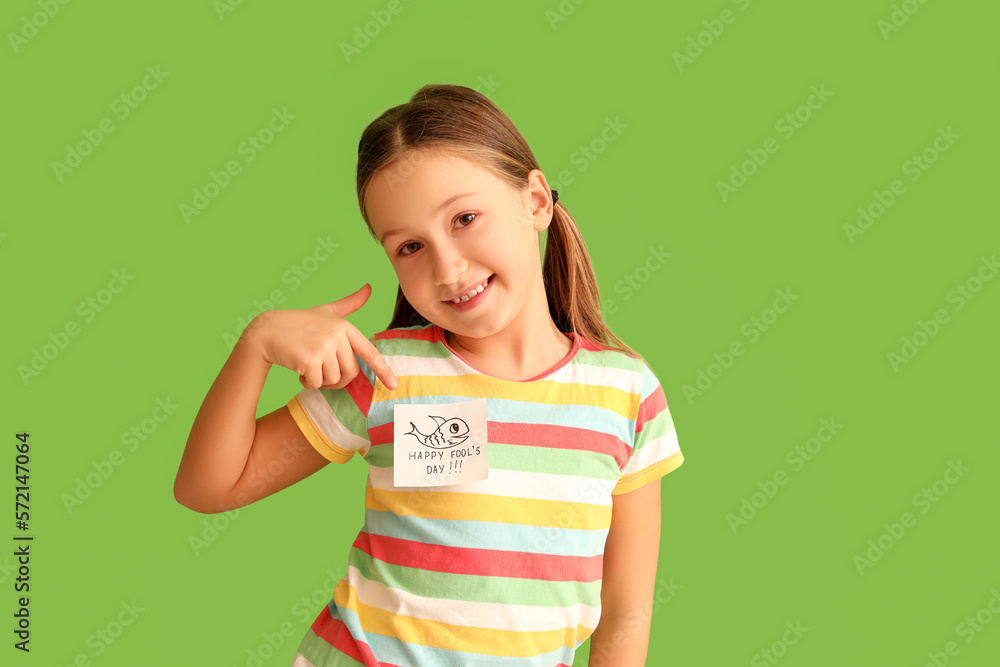 Little girl pointing at sticky paper with text HAPPY FOOLS DAY on her t-shirt against green backgro