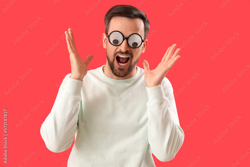 Angry young man in funny eyeglasses on red background. April Fools Day celebration