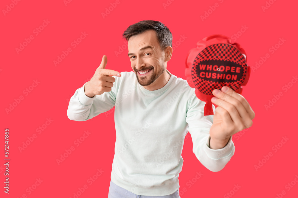 Handsome man pointing at whoopee cushion on red background. April Fools Day celebration