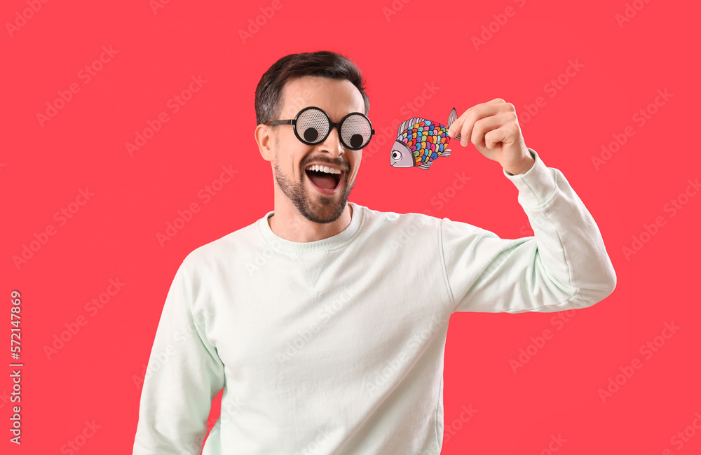 Handsome man in funny eyeglasses with paper fish on red background. April Fools Day celebration