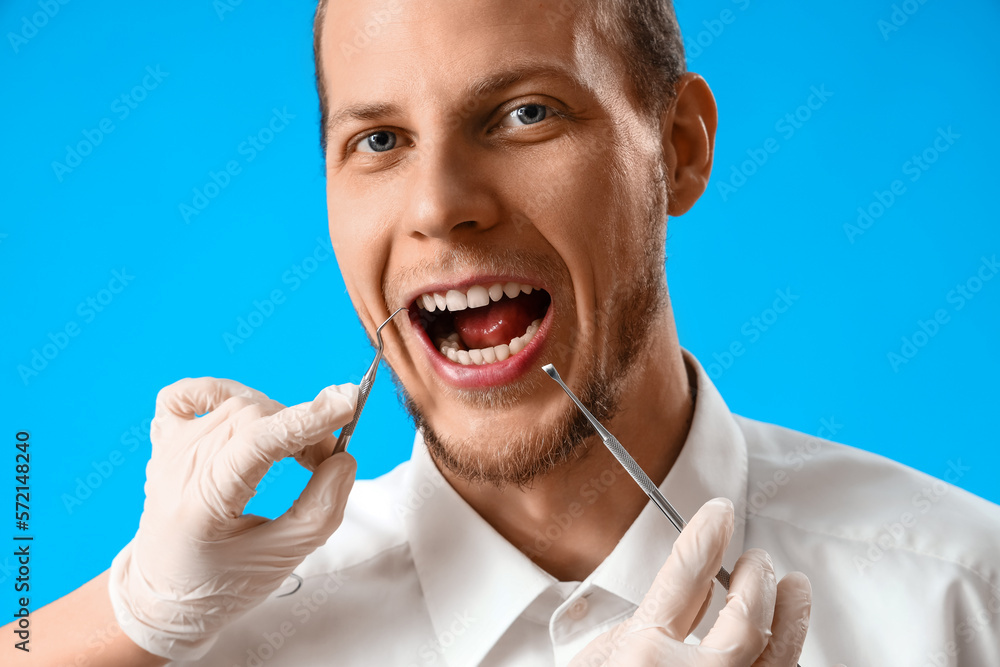 Young bearded man and dentists hands with tools on blue background, closeup