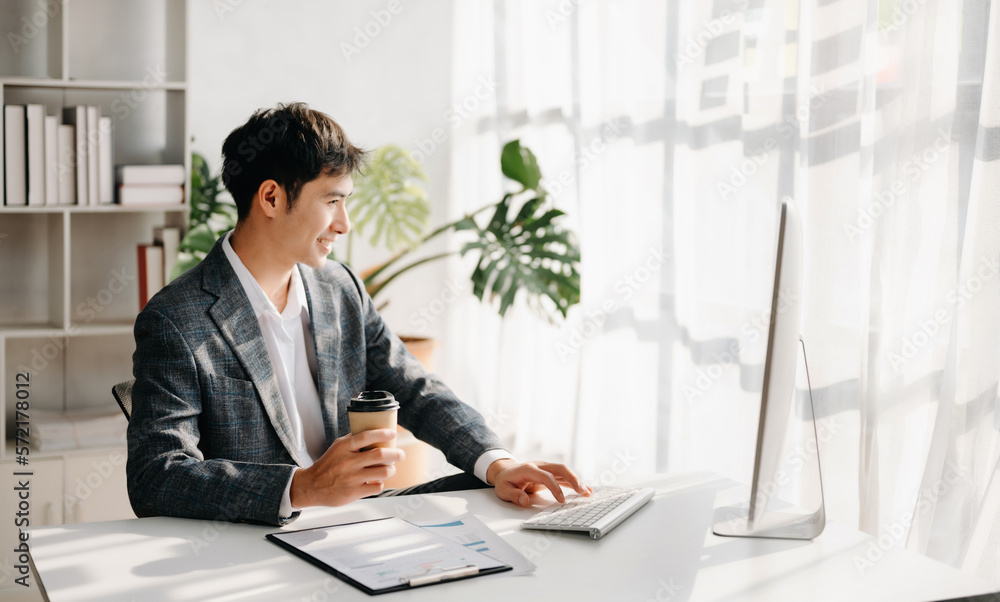 Confident Asian businessman typing laptop computer and digital tablet while holding coffee at modern